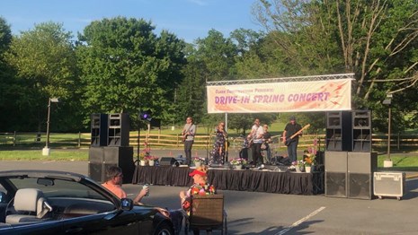 A band performs on a small stage in a parking lot with a banner that reads "Drive In Spring Concert"
