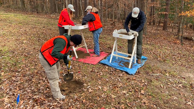 A group of archeologists screening soil from a nearby shovel hole against a winter forest backdrop.