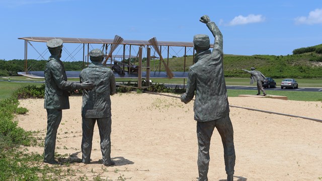 A metal scuplture of three men looking at the Wright brothers flyer on a sandy field