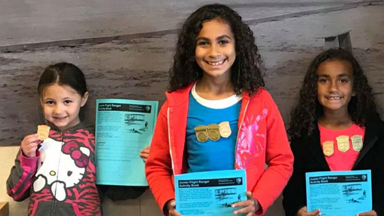 color photo of three girls standing holding blue activity books wearing wooden badges