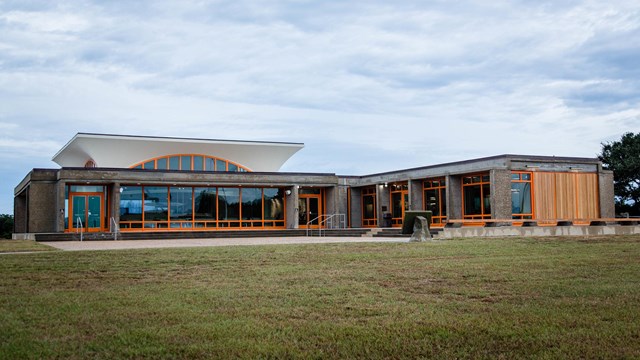 A concrete single-story building with large windows and bright orange window frames in a grass field