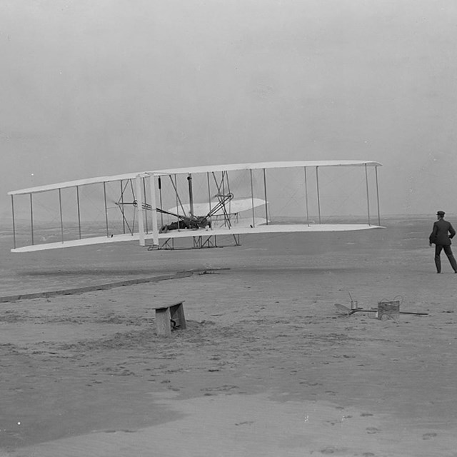 The Wright Flyer is shown in the moment of first takeoff with Orville as pilot and Wilbur alongside