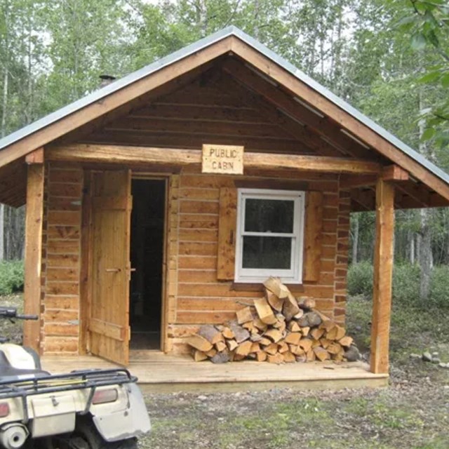 A light brown cabin with a four-wheeled vehicle in a small forest clearing. 