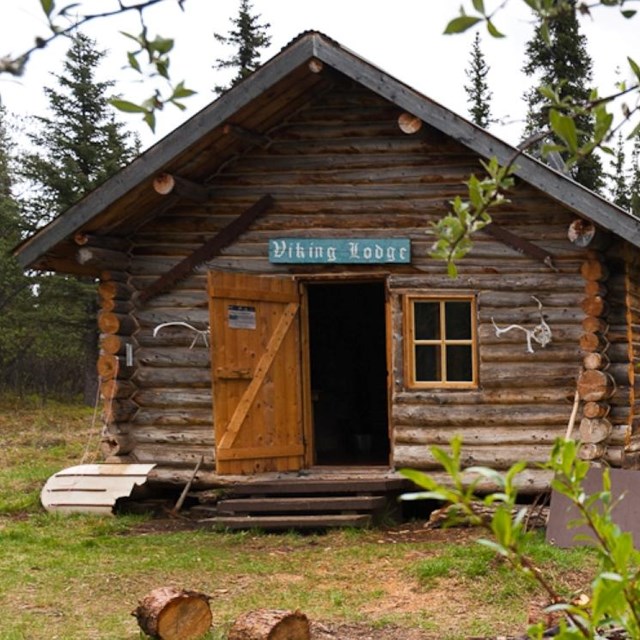 A dark wood cabin sits in a sparse clearing with a small stone pile.
