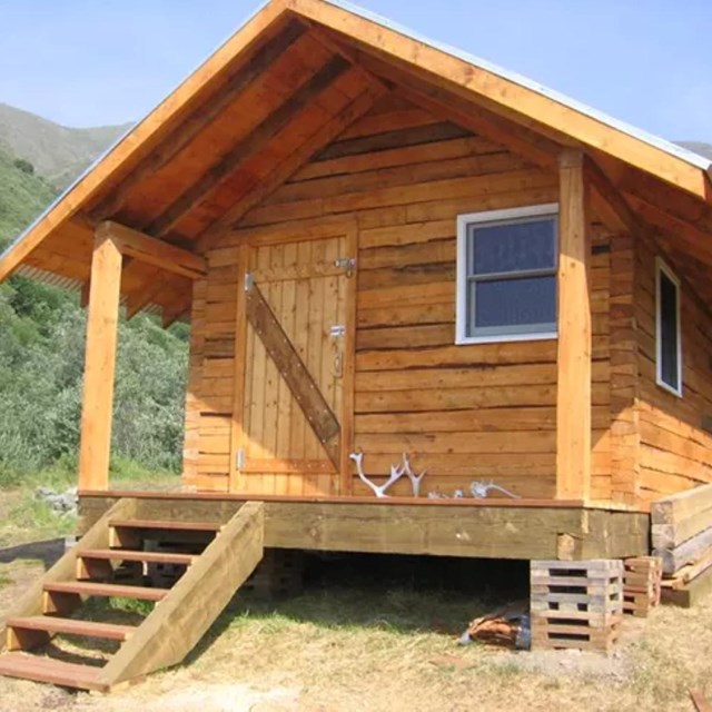 A light wood cabin standing in a high clearing in some hills.