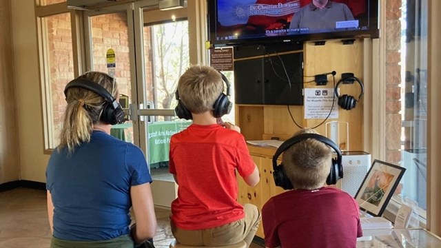 Two young children sit with their mother, all wearing headphones while watching the park film.  