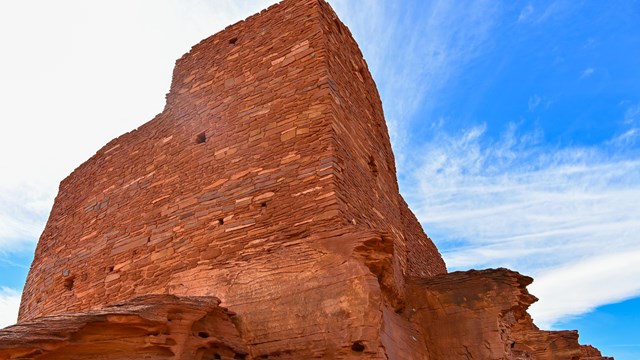 A close-up view of a towering red sandstone pueblo built atop a large matching rock. 
