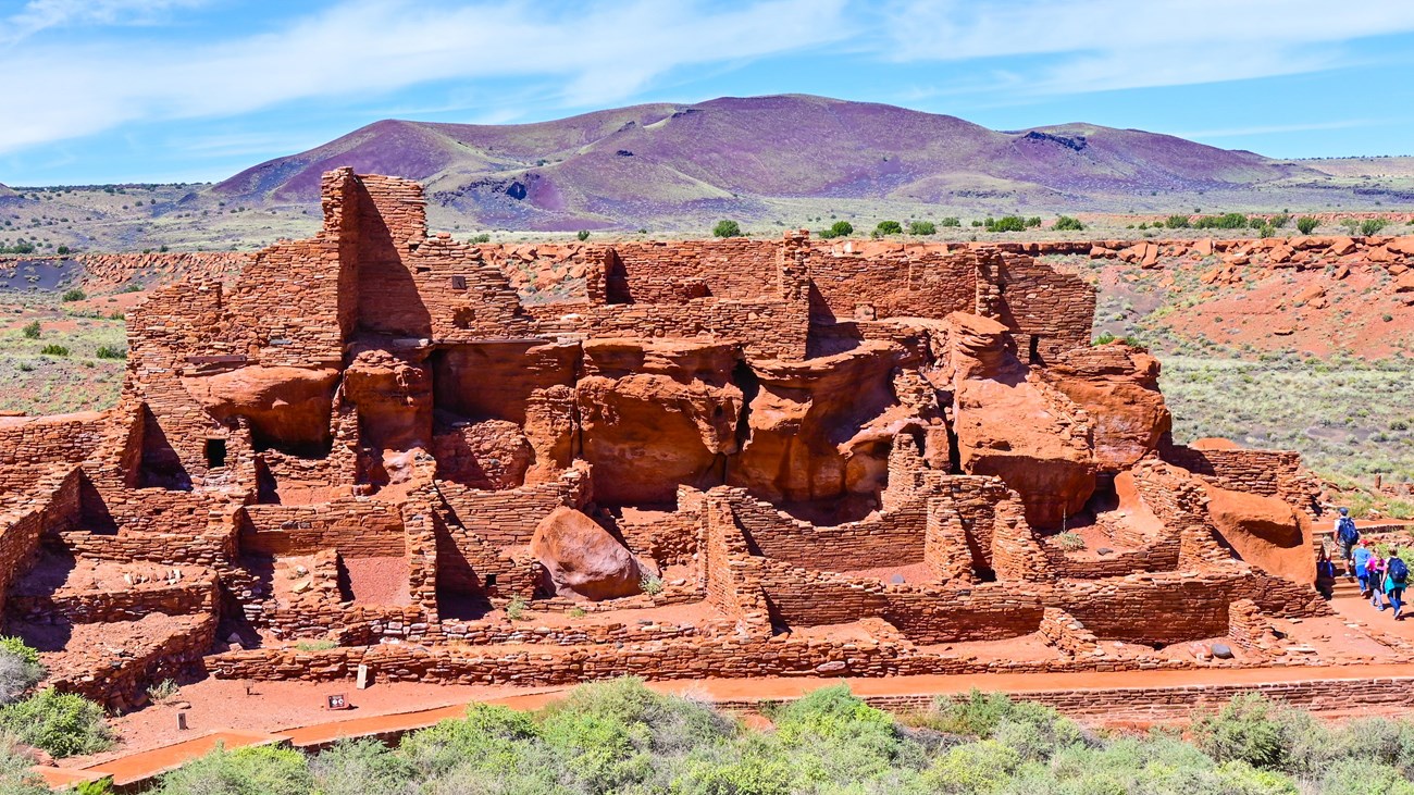 A large red sandstone pueblo with mostly sunny skies and mountains in the background.  