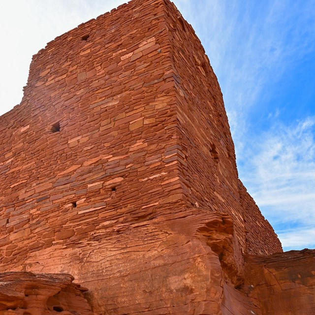 A tall red sandstone pueblo is set against a blue sky with white clouds 