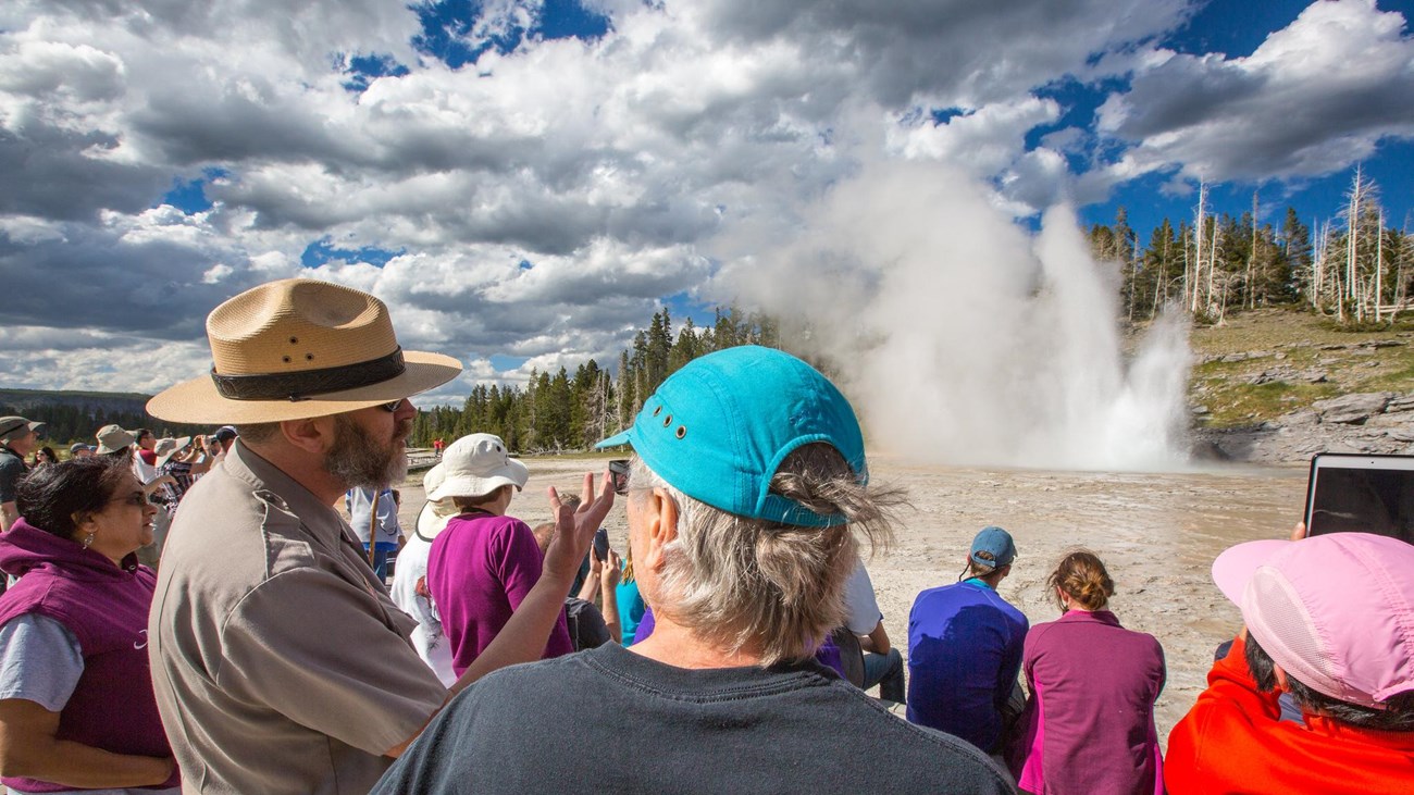 A park ranger speaks to visitors facing an erupting geyser from a crowded boardwalk