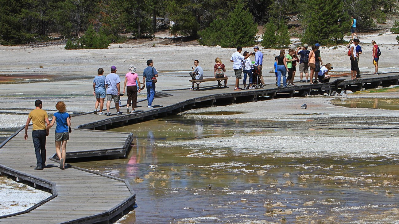 People walk along a boardwalk that goes through the a bare landscape covered in parts by water.