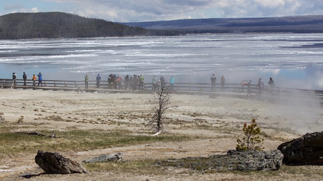 Visitors walk along a boardwalk between hydrothermal features and a lakeshore.