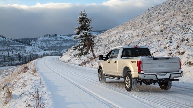 A silver truck is driving down a snow-covered road with snowy mountains in the background.