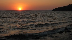 A red and orange sun sets on a large dark body of water.