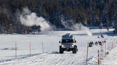 Snowcoach followed by snowmobiles on groomed road with geysers in the background