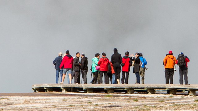 People walk along a board looking a a steam-generating hot spring.