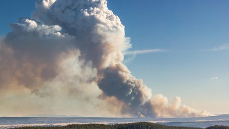 A smoke plume rises into a blue sky across a wide landscape