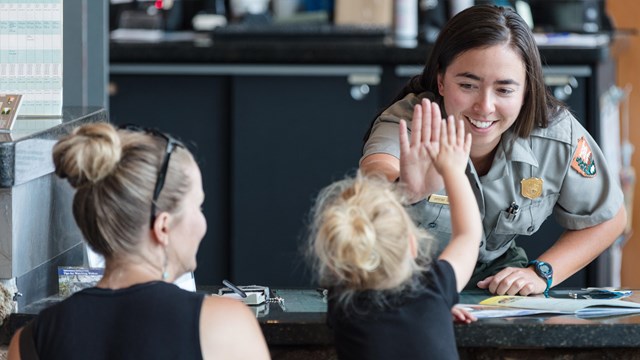 A child with a woman gives a high 5 to a park ranger across a desk.