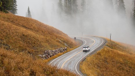 A car drives along on a winding road during a foggy morning.