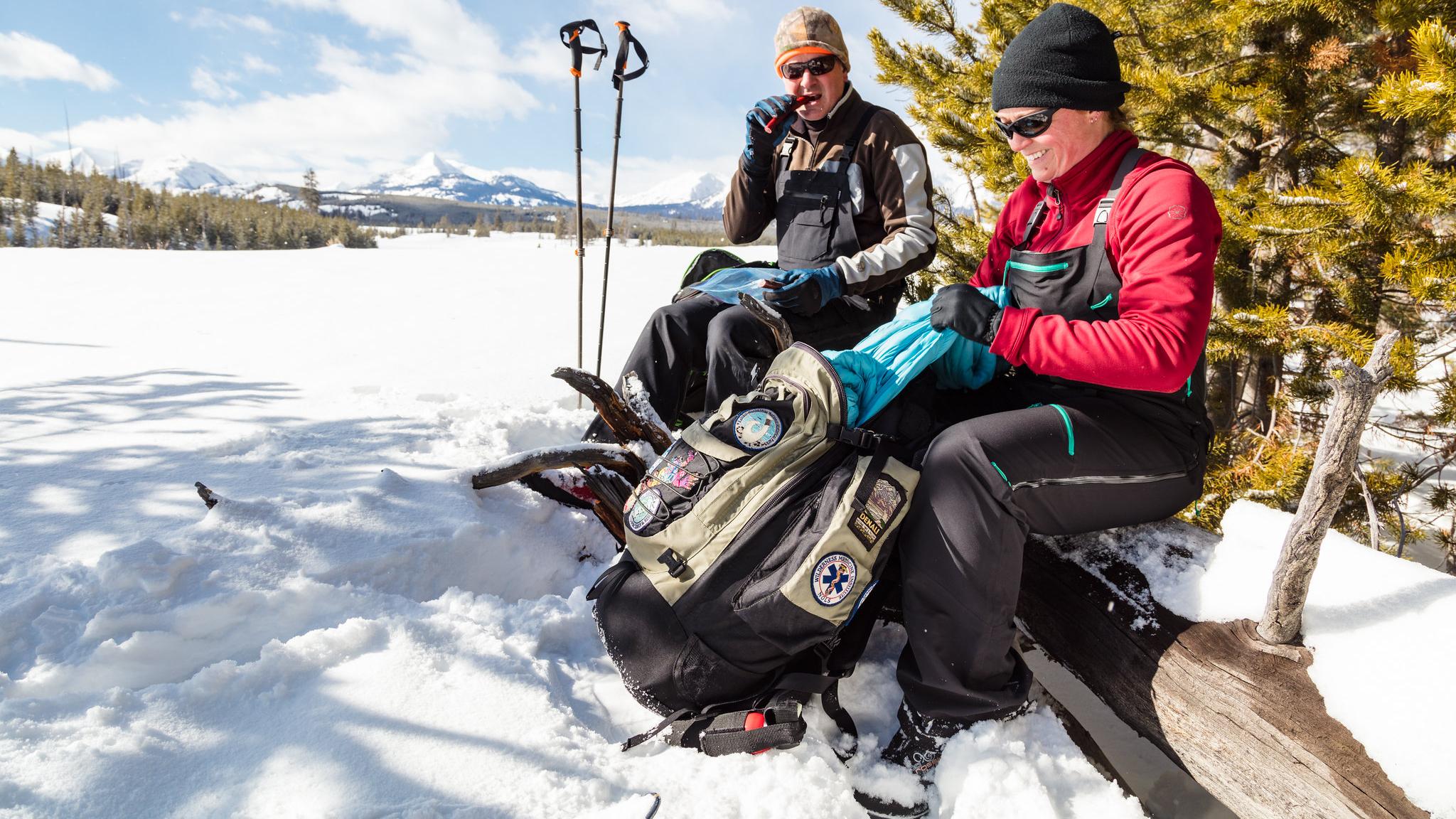 A pair of skiers on change layers and eat while out for a ski.