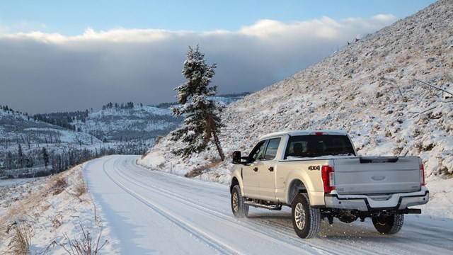 A truck drives on a layer of snow on a paved road with a snow-covered mountain in the background.