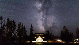 The milky way in the night sky rises above a lighted building, surrounded by tall trees.