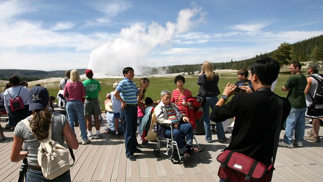 Wheelchair-bound visitor and his family take a picture while Old Faithful Geyser erupts.
