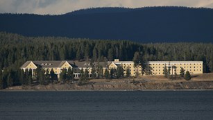 Bright yellow, three-story hotel building standing in the conifer forest next to a deep, blue lake.