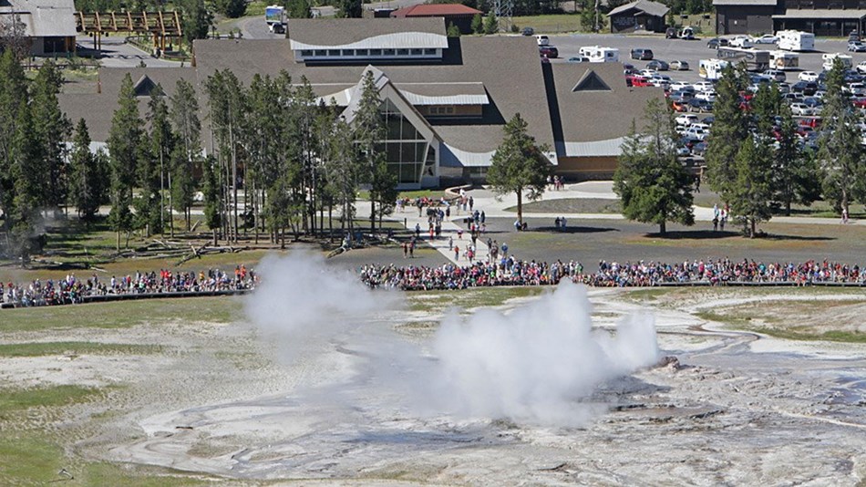 Steam rolls up from a vent with visitors watching along a boardwalk and buildings behind.