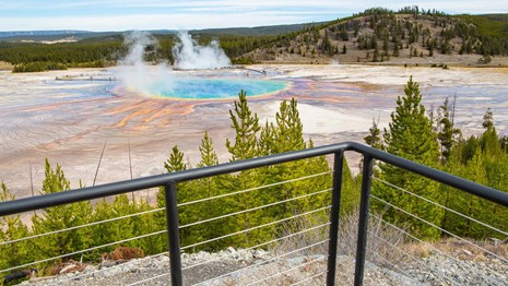 A metal and cable railing in front of an overlook of a blue pool surrounded by prismatic colors.