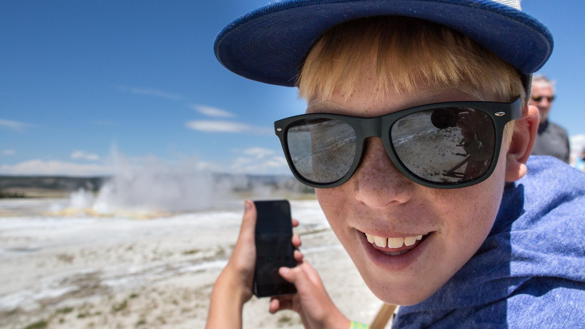Close up of a young boy with a hat and sunglasses taking a picture of a geyser with a phone.