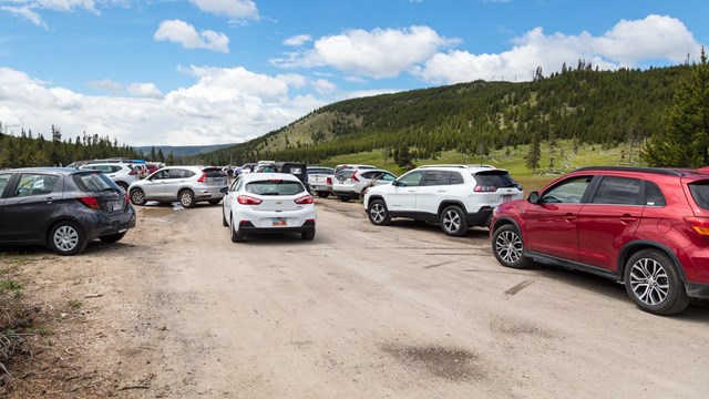 Cars line the bare ground parking area and other cars wait for a parking space.