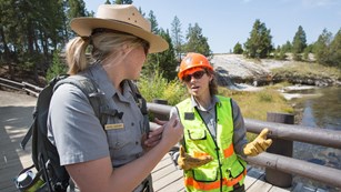 Ranger wearing a yellow safety vest talks with another ranger while standing on a bridge.