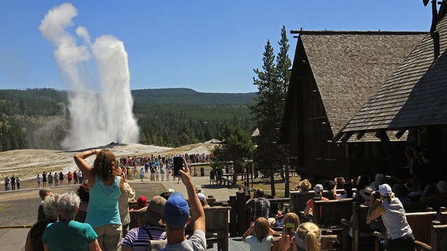 People sitting and standing on a wooden patio taking pictures of an erupting geyser.