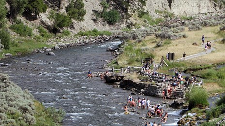 Brightly-clothed people in a river near a steaming thermal feature