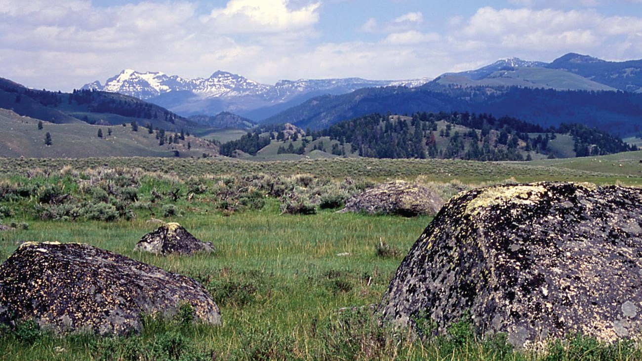 A trail leads to a boulder on top of a hill with snow-capped mountains