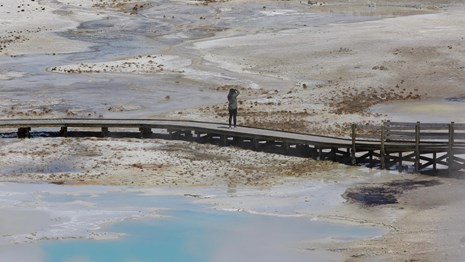 A lone person standing on a boardwalk and takes a picture of steaming hot springs.
