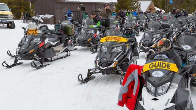 Three lines of snowmobiles wait for their drivers to return.