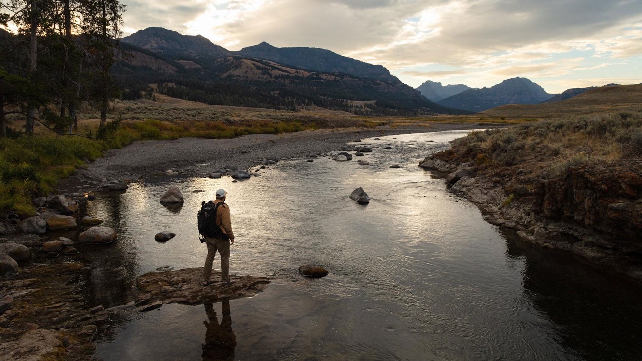 A hiker stands on a rock on the edge of a river in the fading daylight.
