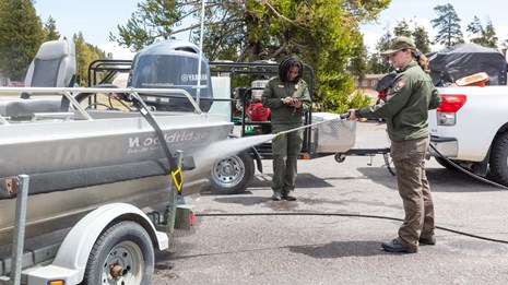 One park ranger sprays a motorboat with a hose, while another ranger writes on a clipboard nearby.