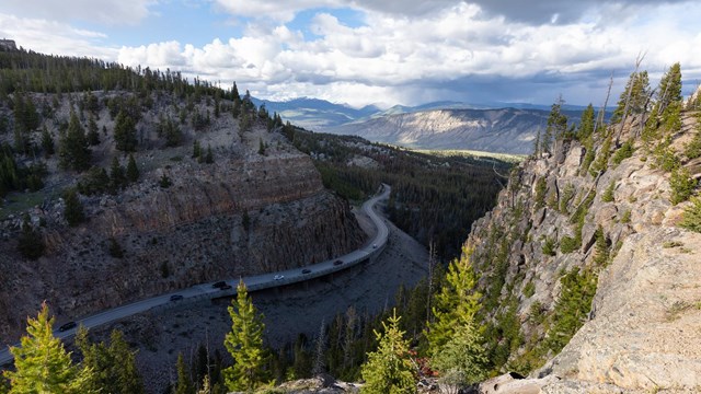 Cars drive on a bridge that hugs the edge of a large cliff through the mountains.