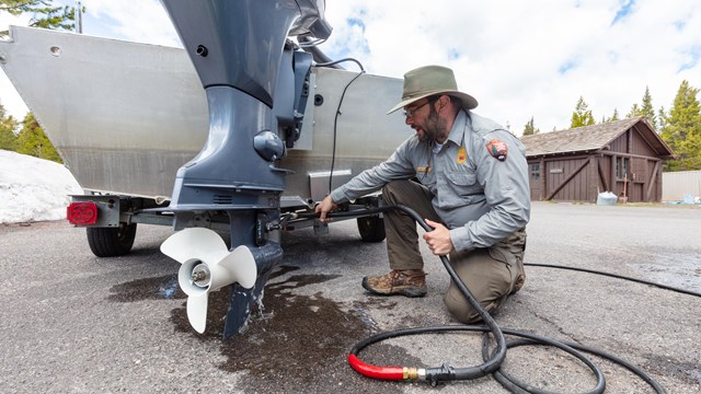 A park ranger with a dark beard and wearing a green and grey uniform kneels with a hose next a boat.