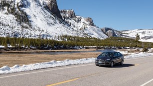 A car drives along a road next to a river, with snow still covering the nearby mountain slopes.