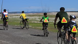 A family pedals by some thermal features along a paved path on bicycles.