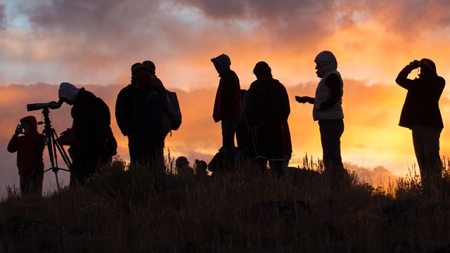 Silhouette of people taking photographs and watching through spotting scopes at a colorful sunset.