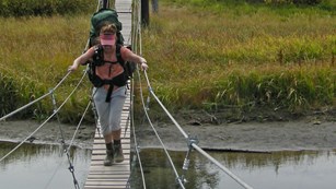 A woman with a large backpack carefully crosses a small suspension bridge.