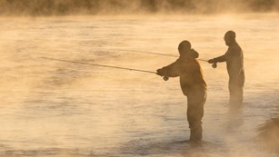 Two anglers stand in a river and cast their lines during an early morning sunrise.
