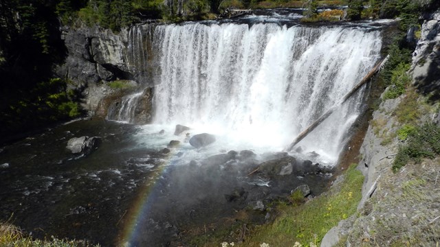 A waterfall drops over a gray rock cliff and a rainbow glows in the spray.