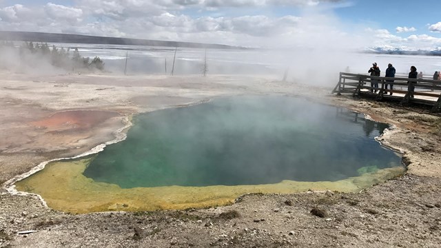 Visitors stand on boardwalk and gaze into a deep, blue pool rimmed in yellow and orange.