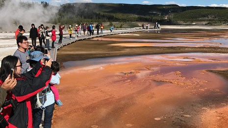 People line the boardwalk, looking out and taking pictures of a steaming hot spring.
